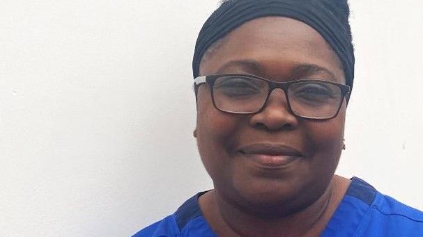 A head and shoulders shot of Eunice Vial. She is wearing a blue NHS branded gown and glasses.