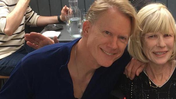 Prof Portmann smiling wearing a navy shirt sitting beside his cousin Anne who has short blonde hair and is wearing a black top.