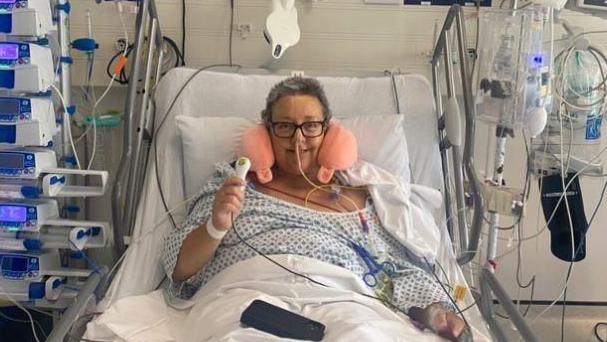 Victoria is in a hospital bed, surrounded by wires, drips and tubes. She has an inflatable neck cushion and is sat up smiling after her surgery.