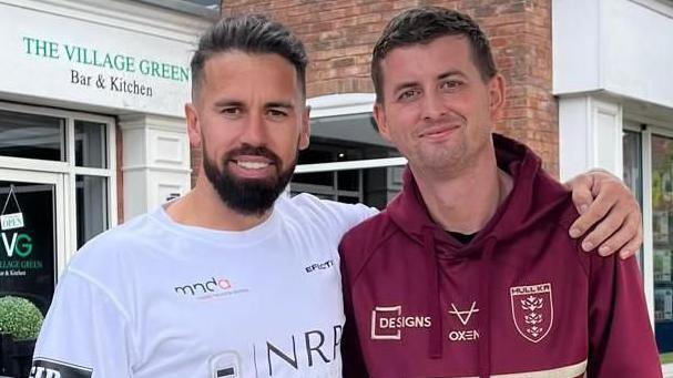 Mike is stood with his left arm around the shoulder of his friend Craig Eskrett. Mike is wearing a white T-shirt with company names and logos on acting as sponsors. He has a black goatee beard and swept-back hair. Craig has a burgundy Hull KR hoodie and has short dark hair. The pair are stood in front of a row of shops and looking into the camera