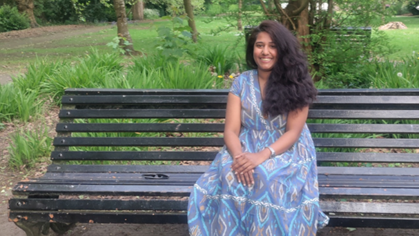 A woman with brown hair and wearing a blue dress sat on a park bench.