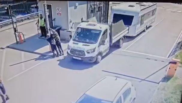 A white tipper truck towing a caravan passes a gatehouse as two men hold up the barrier. A security guard in a yellow jacket stands on the stop of his gatehouse.