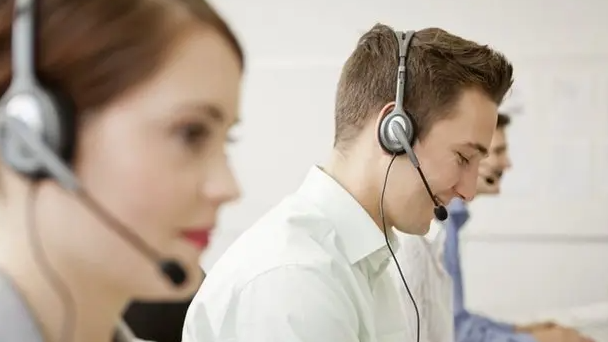 A man and a woman sit in an office with headsets on. The man is smiling and the woman, who has red hair, is also smiling.