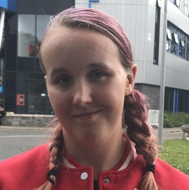 A picture of Abigail in a red jacket. She has pink hair and is stood in front of the college building.