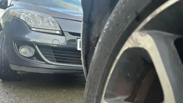 A close-up of two cars parked on a street. The shot has been taken from a low angle. The tyre of one car, which is all that can be seen of it, is in the foreground on the right. The bonnet of another car can be seen on the left of the picture, inches behind the first car.