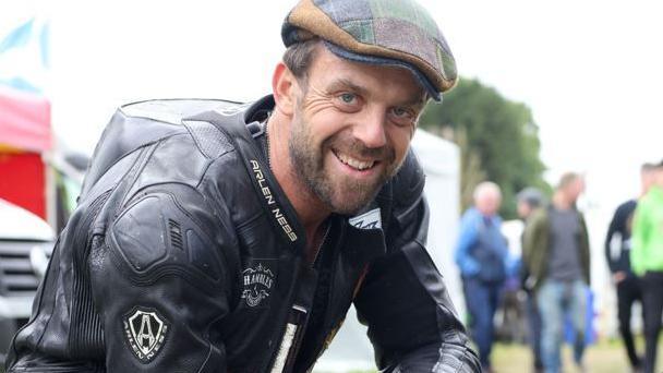 Andy McAllister crouching down wearing black racing leathers and a chequered tartan flat cap. He has short brown hair and a close-cropped beard and is smiling.