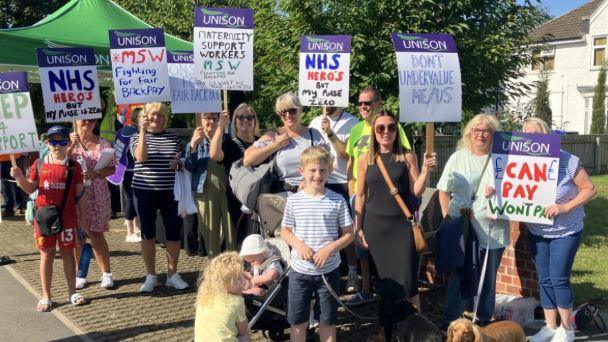 Maternity staff wearing casual clothes, some with children, holding placards with handwritten slogans including "can pay, won't pay" and "don't undervalue me/us".