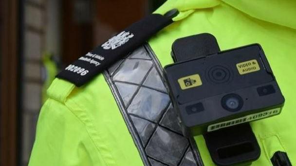 A close up shot of a traffic warden's shoulder, wearing a yellow hi-vis jacket with a small black square camera pinned to the chest, a yellow label on the camera says 'video& audio'