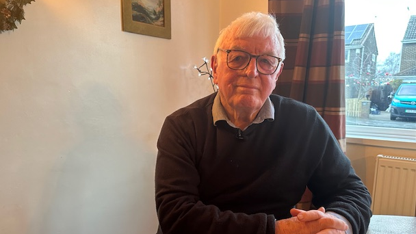 Keith Bell smiling into the camera. He has got white hair and is wearing glasses. He is sitting in front of a window with tartan curtains.