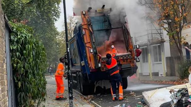 Refuse workers use hand held fire extinguishers to try and put the fire out in the back of a bin lorry. A large amount of smoke and some flames can be seen