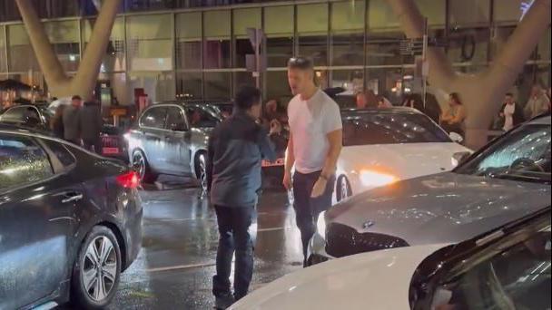 A man in a white t-shirt and jeans with sunglasses head stands face to face with a smaller man in a grey jack in the middle of a road on a rainy evening. The two can be seen looking animated, while cars idle all around them. 