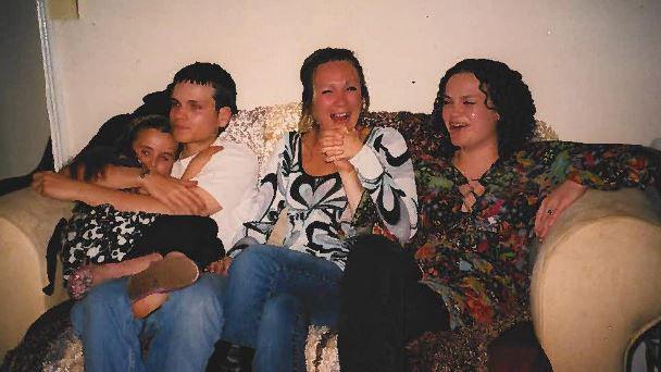 Lewis Skelton sitting with his sisters on a sofa with his two older sisters holding hands and laughing and Lewis cuddling his younger sister