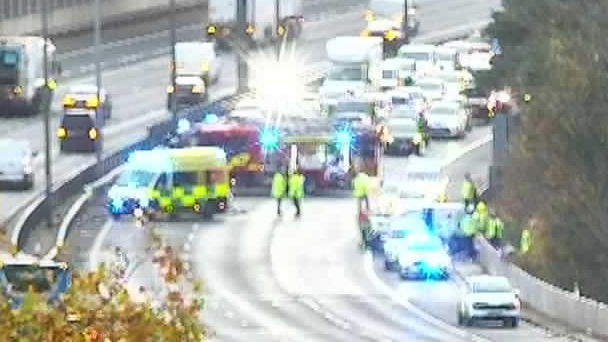 A blurry camera image of a crash on the M40. An ambulance is parked on the right in the outside lane; a few emergency workers are standing in the middle lane and nearside lane. Behind it is a e fire engine sideways on blocking all three lanes with queuing traffic behind.