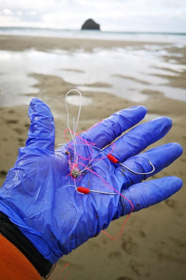 A hand wearing a plastic glove holds two barbed fishing hooks threaded with fishing line. The person is standing on the beach with the sea and a rock in the distance.