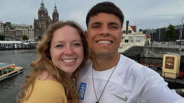 A man wearing a white football top smiling with his arm around a woman wearing a yellow top. Behind them is a city scene.