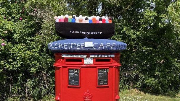 A knitted postbox topper features a boat and several knitted faceless figures. The topper says Alzheimer Cafe on the side.
