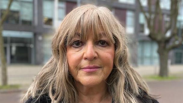 Nadia Cenci smiles at the camera. She has shoulder length blonde hair and a fringe. The background, consisting of buildings and trees, is blurred.