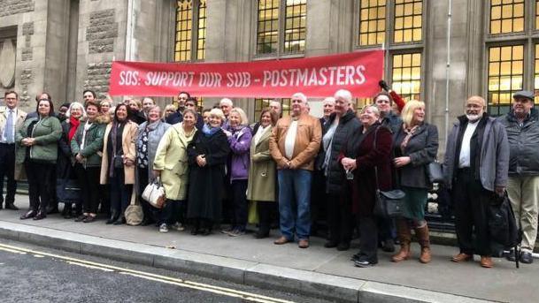 A crowd of people - mainly middle-aged - standing in front of a stone building with a red banner above their heads, which reads: 'SOS: SUPPORT OUR SUB-POSTMASTERS'.