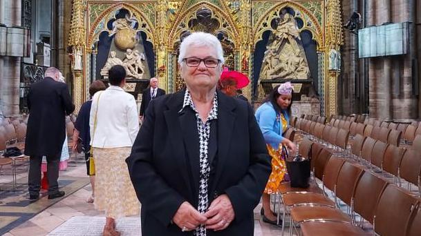 Margaret Peacock at King Charles' coronation in 2022.  She has short white hair and glasses and is wearing a long black coat over a black and white blouse.