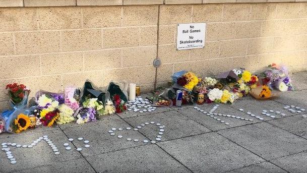 Bunches of flowers and candles are placed against a wall. Cans of drink are left too. Tealights spell out the names Mo and King with a heart depicted by tealights. 