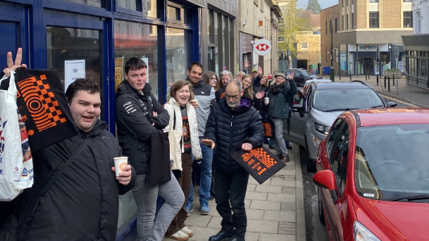 On George Street, people queue on the left-hand side of the pavement in single file. Two people have bags. People are smiling and looking at the camera. 