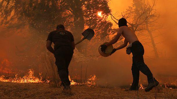 Two men stand in front of a flaming tree. One throws water on the flames from a bucket