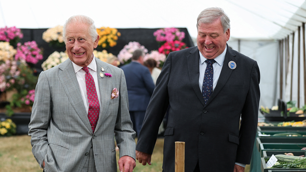 King Charles III walks alongside Sandringham Flower Show chairman Graham Browne. The King is wearing a grey suit and is smiling. Graham is wearing a black suit and is smiling his gaze at the floor.