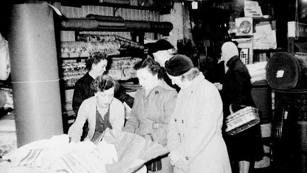 An old black and white image of Joe Elphick's daughter, Brenda, serving customers. There are three women at the front of an image looking down at some material. 