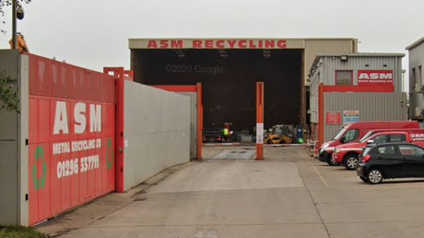 The outside of ASM Metal Recycling at its site on Griffin Lane in Aylesbury showing a large open-sided warehouse. There is a car and two vans parked outside.