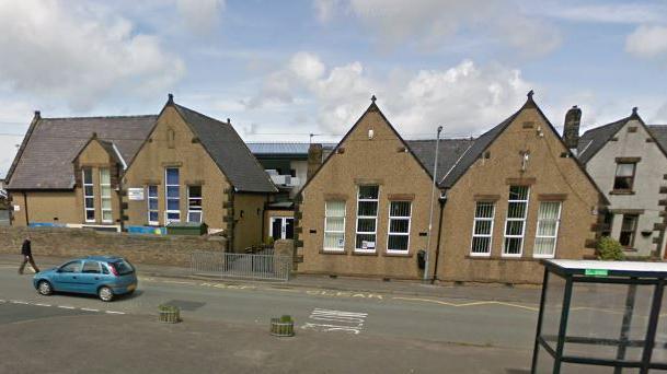 A Google Street View screenshot of Broughton Moor Primary School. The building is off the main street through the village and is covered in brown pebbledash. It has long and narrow windows on each section of the building.