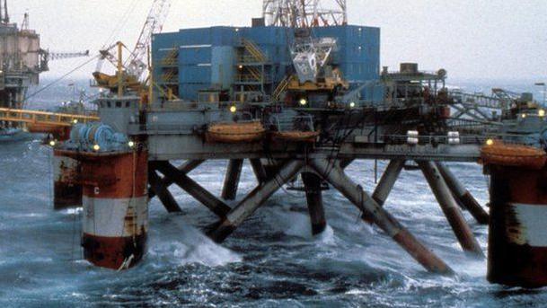 A large floating platform in a sea with five giant red and white legs, a lot of metal work and three tiers of blue storage containers on its deck which have been converted into accommodation.