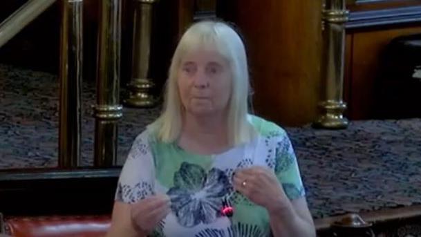 A woman with white blonde hair and a fringe is smiling. She is wearing a white top with green spots and black flowers on it. 