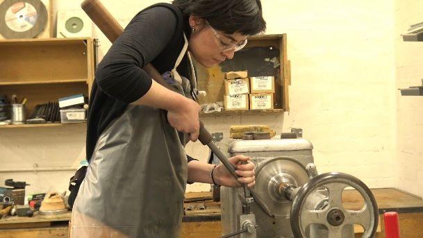 Lily Smith, in her workshop, leans over her workbench where her silver spinning maching is turning. She is wearing an apron and protective goggles while she holds a long steel stick against the turning parts. Wall shelves with various boxes, tools and pieces of equipment are behind her as she concentrates.