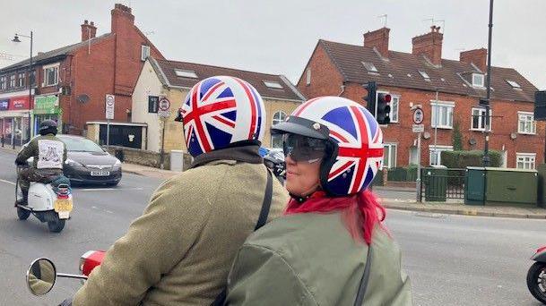 A man and a woman ride a scooter driving down the road wearing green coats and Union Jack helmets