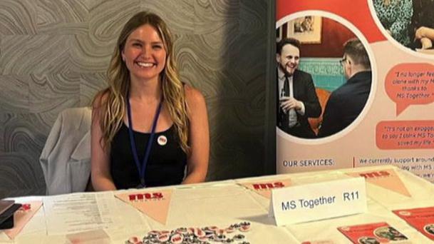 Amy is sitting on a chair next to an advertising sign for her charity. It reads: "MS Together. Our charity provides services of advice and support to people people affected by multiple sclerosis (MS) with a particular focus on those aged 18-35 who are living with MS."