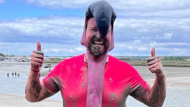 Jim Rugg with a big smile on his face and giving a double thumbs up. He is wearing a muddy pink flamingo outfit with a large black beak drooping over his face. Behind him is the muddy river through which he has just ran.
