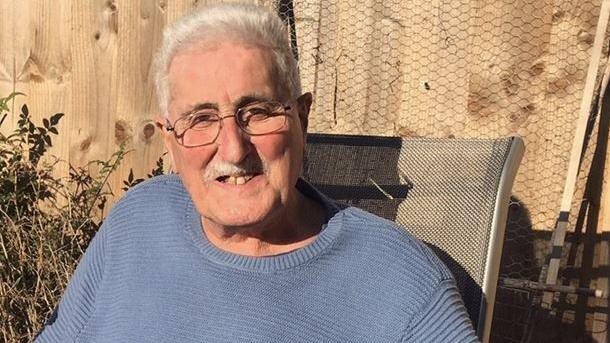 A smiling man with white hair and a white moustache and glasses sits in the garden in the sun. A tanalised timber fence and some shrubbery is visible behind him.