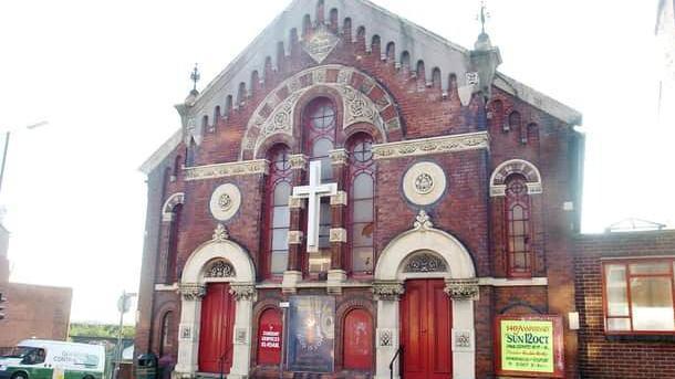 A Victorian church with a sloping roof, a white cross front and centre of the building and matching sets of red doors.