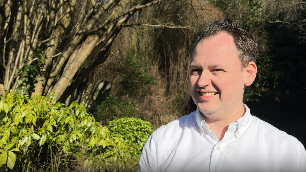 Close up of Scott Bloomfield who is standing outside next to a road. Trees and bushes are behind him. Mr Bloomfield is wearing a white shirt and is smiling at the camera. 