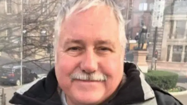 James Cochrane looking straight at the camera. He has white and grey hair and a white an grey moustache and is wearing a dark jacket.