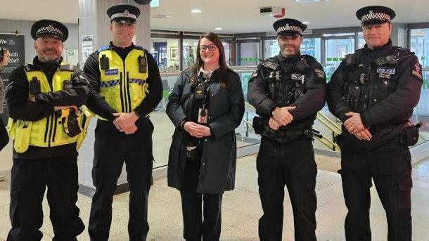 Four police officers in uniform - two from Devon and Cornwall Police and two from British Transport Police - stand either side of a woman inside a train station