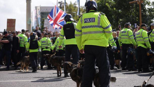 A police dog handler in riot gear and his police dog stand alongside other handlers and dogs and a row of police officers in bright green jackets facing a a group of shouting men and women waving Union Jacks and St George flags