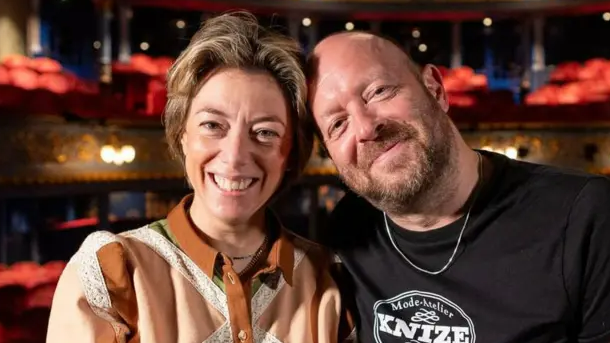 Nicole Taylor, with short highlighted brown hair and wearing a brown and peach shirt, and John Tiffany, a bald man with a short beard wearing a black t-shirt with a Knize logo, sit together smiling at the Lyceum Theatre in Edinburgh.