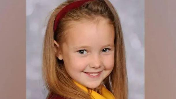 Elsie Dot Stancombe wears a red hairband and school uniform and smiles at the camera