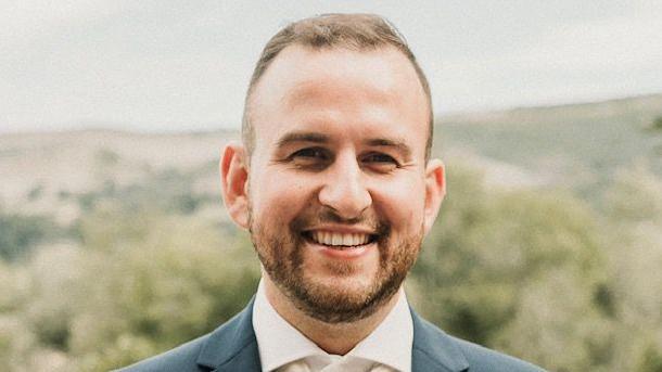 Alun Owen facing the camera and smiling, with a white formal shirt and petrol blue suit jacket