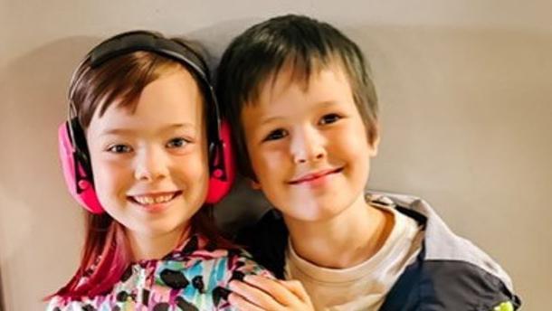 A young girl with brown hair and a patterned top has pink ear defenders on, and a boy with brown hair and a white T-shirt and black jacket has his arm around her. They are both smiling at the camera.