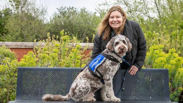 Anne Herbert is standing behind a park bench. Her support dog Rafa is sitting in front of her on the bench. He is a labradoodle with grey and black markings.