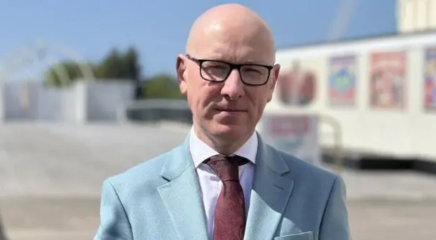 Patrick Hurley has a bald head, dark spectacles, a white shirt, maroon tie and sky blue jacket