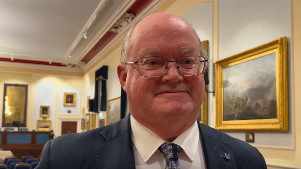 Constable Simon Crowcroft smiles at the camera. He is wearing a blue suit with a white shirt and patterned blue tie. He is in a meeting hall. He is wearing glasses.