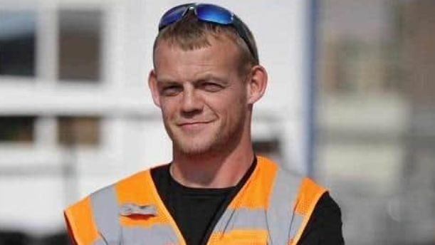 Andrew Main, smiling into the camera. He is wearing an orange hi-vis jacket and has sun glasses resting on his head. 